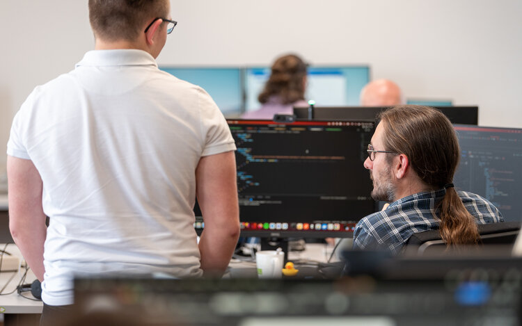 2 Männer im Vordergrund sitzen mit dem Rücken zum Betrachter vor einem Monitor